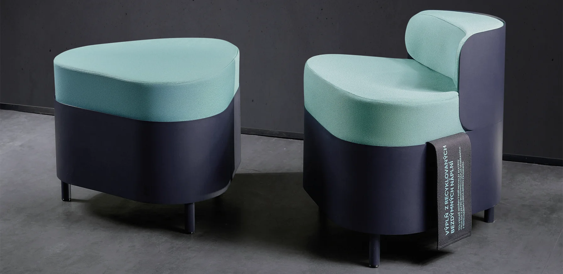 Two modular lounge seats with mint green upholstery and dark bases, one featuring an informational sleeve about recycled filling, set against a grey brick wall.
