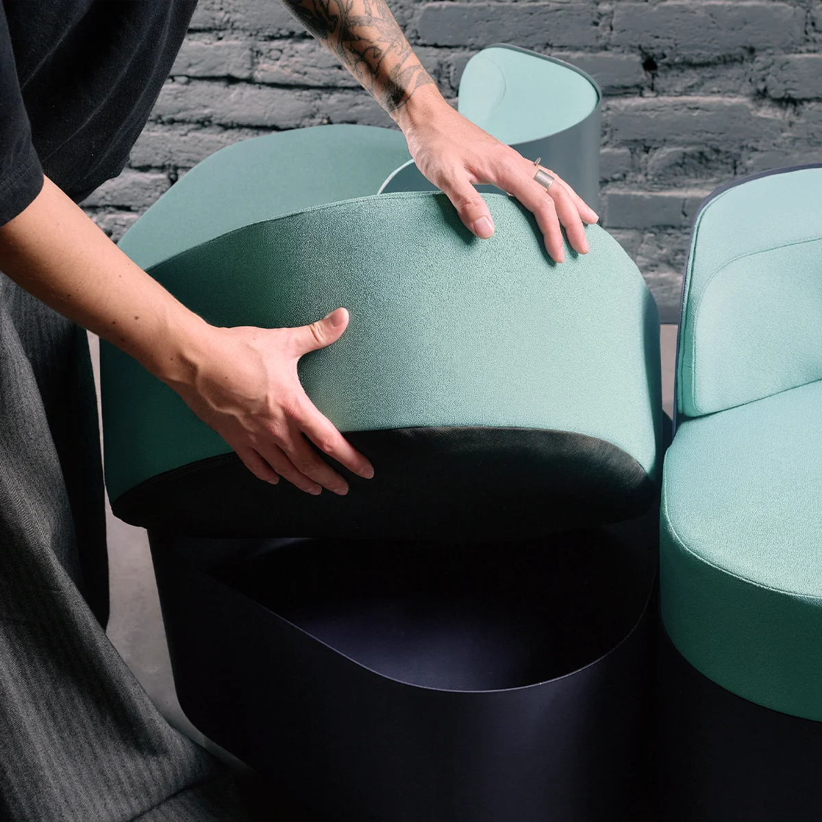 Close-up of a person lifting the seat cushion from a modular lounge chair, revealing the hollow interior. The chair features a curved teal cushion and a dark base.