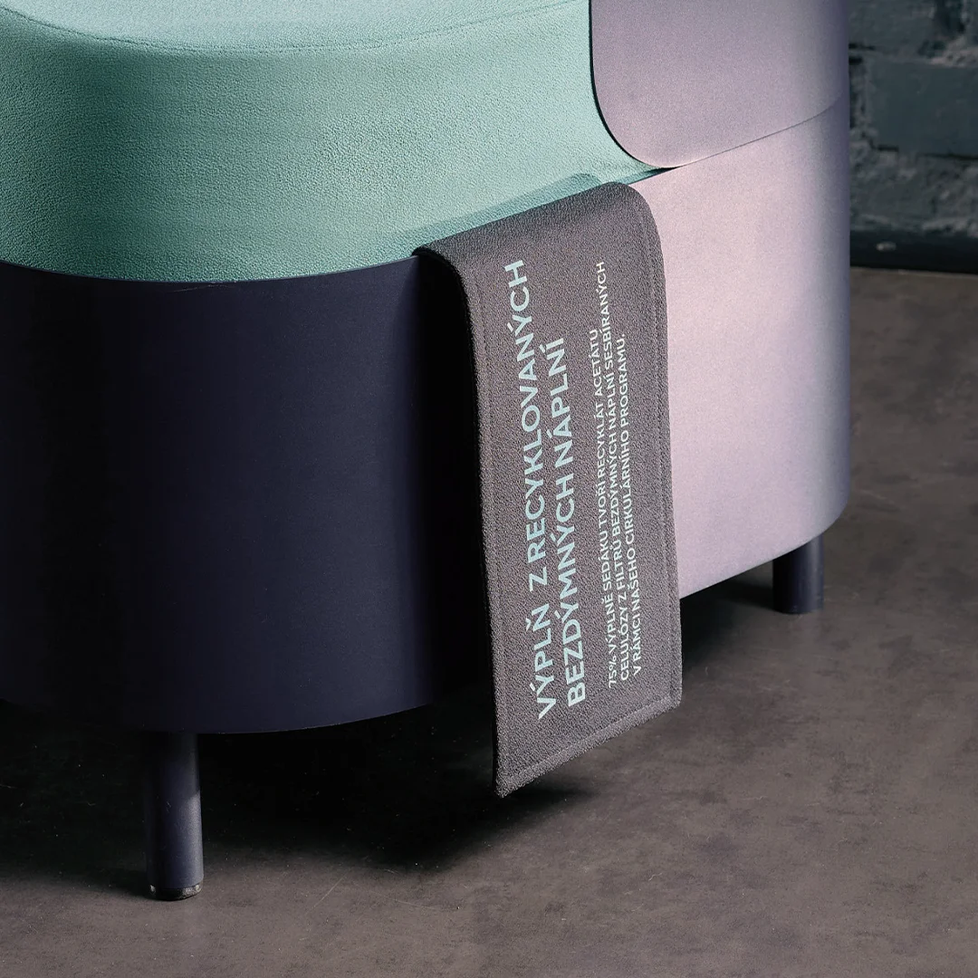 Close-up of a modular lounge chair showing a turquoise upholstered seat and dark navy base. A black woven label is attached, printed with Czech text indicating that the chair’s filling is made from recycled tobacco stick filters.