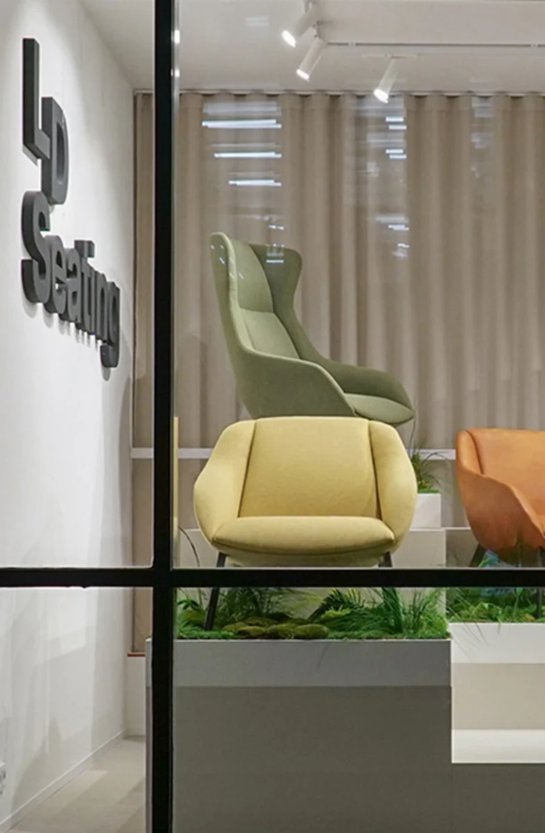 LD Seating showroom window displaying Terra Lounge armchairs in soft colours, arranged on tiered platforms with greenery, illuminated at night and viewed from the street.