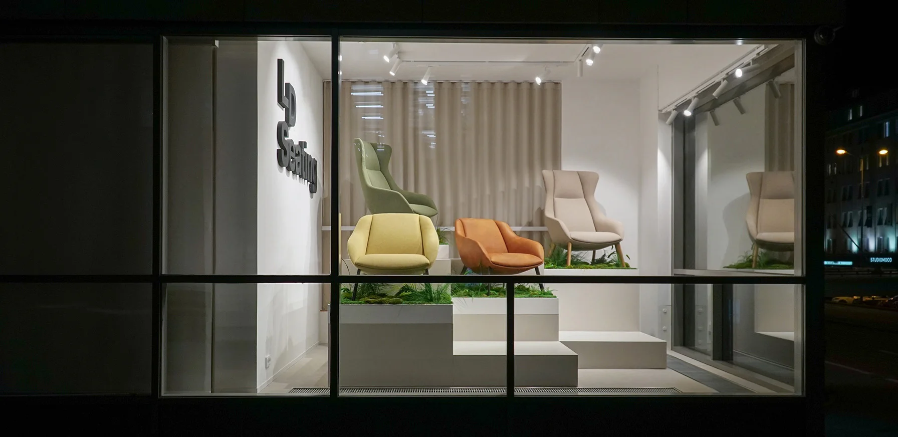 LD Seating showroom window displaying Terra Lounge armchairs in soft colours, arranged on tiered platforms with greenery, illuminated at night and viewed from the street.