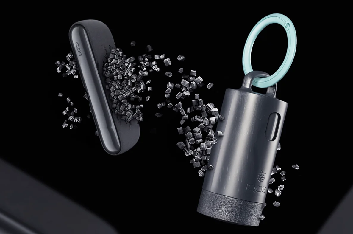 IQOS Clip Case with a turquoise carabiner shown among recycled plastic pellets and IQOS devices on a dark background.